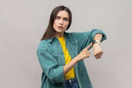 Téléchargez les photos : Concerned punctual dark haired woman pointing finger at smartwatch on her wrist, look at time, hurry up and act, wearing casual style jacket. Indoor studio shot isolated on gray background. - en image libre de droit