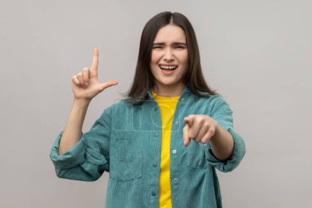 Téléchargez les photos : Disrespectful rude young woman showing loser gesture and pointing on you, abuser, mocking your failures, wearing casual style jacket. Indoor studio shot isolated on gray background. - en image libre de droit
