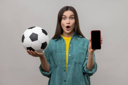 Téléchargez les photos : Shocked surprised woman with dark hair holding and showing blank display smartphone and football ball, betting on soccer, wearing casual style jacket. Indoor studio shot isolated on gray background. - en image libre de droit