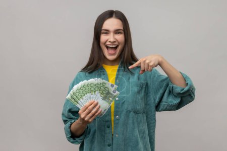 Foto de Delighted woman being happy to hold big money, pointing to euro bills in hand and looking with positive excited expression, wearing casual style jacket. Indoor studio shot isolated on gray background. - Imagen libre de derechos