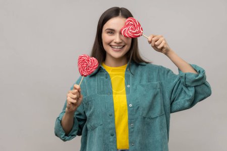 Foto de Childish woman with dark hair covering eye with heart shape lollipops, hiding, having fun with sweets, wearing casual style jacket. Indoor studio shot isolated on gray background. - Imagen libre de derechos