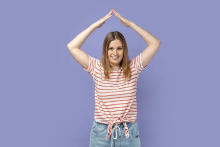 Téléchargez les photos : I'm in safety. Portrait of charming positive blond woman wearing striped T-shirt gesturing house roof symbol over head, feeling protected. Indoor studio shot isolated on purple background. - en image libre de droit