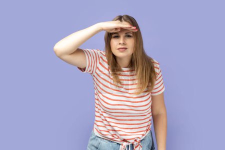Photo for Portrait of attentive beautiful blond woman wearing striped T-shirt standing with hand near forehead and looking far away, looks at distance. Indoor studio shot isolated on purple background. - Royalty Free Image