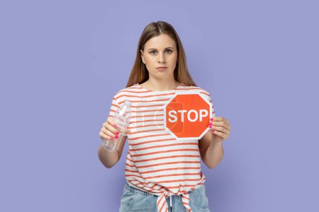 Foto de Portrait of serious strict bossy young blond woman wearing striped T-shirt showing red stop sign and holing plastic bottle in hands. Indoor studio shot isolated on purple background. - Imagen libre de derechos