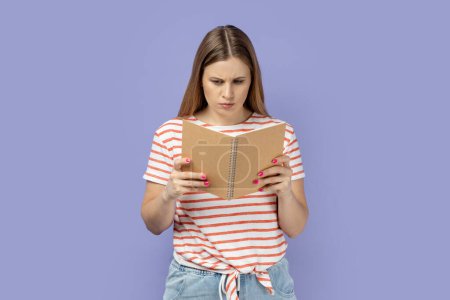 Foto de Portrait of serious concentrated blond woman wearing striped T-shirt holding and reading book, being impressed by plot, reads with attention. Indoor studio shot isolated on purple background. - Imagen libre de derechos