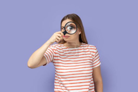 Foto de Portrait of funny positive blond woman wearing striped T-shirt standing, holding magnifying glass and looking at camera with big zoom eye. Indoor studio shot isolated on purple background. - Imagen libre de derechos