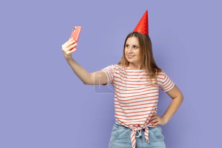 Téléchargez les photos : Portrait of blond woman blogger wearing striped T-shirt and party cone broadcasting livestream or taking selfie for her blog, keeps hand on hip. Indoor studio shot isolated on purple background. - en image libre de droit