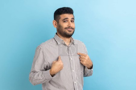 Portrait of bearded businessman pointing himself and looking selfish egoistic haughty, feeling proud of own achievement, wearing striped shirt. Indoor studio shot isolated on blue background.