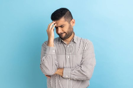 Téléchargez les photos : Businessman with beard making facepalm gesture keeping hand on head, blaming himself for bad memory, unforgivable mistake, wearing striped shirt. Indoor studio shot isolated on blue background. - en image libre de droit