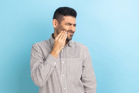 Photo for Unhappy bearded businessman feeling toothache, touching sore cheek, suffering from cavities, cracked teeth, gum recession, wearing striped shirt. Indoor studio shot isolated on blue background. - Royalty Free Image
