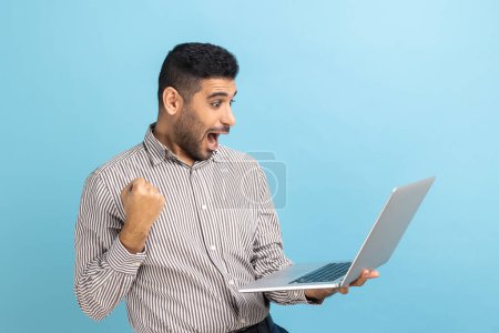 Téléchargez les photos : Excited businessman with beard screaming yes raising hands up, solving hard task, getting access, successfully completing work, wearing striped shirt. Indoor studio shot isolated on blue background. - en image libre de droit