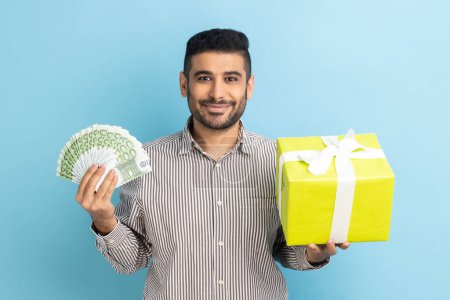 Photo for Buying holiday gifts. Bearded businessman holding wrapped present box and money euro, looking at camera smiling satisfied, wearing striped shirt. Indoor studio shot isolated on blue background. - Royalty Free Image