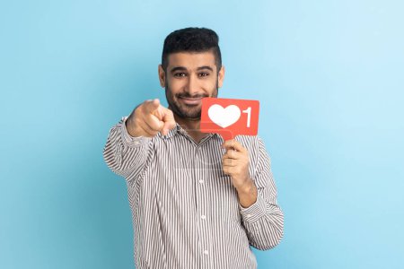 Téléchargez les photos : Do you rate my post in social network? Smiling man blogger holding like counter sign and pointing finger at camera, wearing striped shirt. Indoor studio shot isolated on blue background. - en image libre de droit