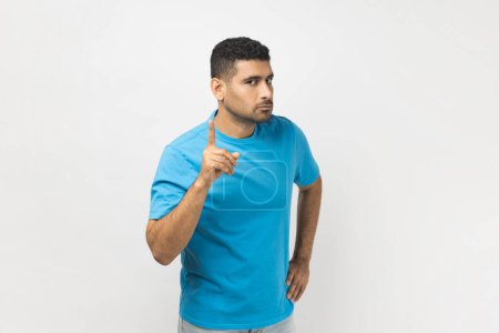 Foto de Portrait of serious bossy attractive unshaven man wearing blue T- shirt standing with raised index finger, warning somebody, showing disapproval gesture. Indoor studio shot isolated on gray background - Imagen libre de derechos