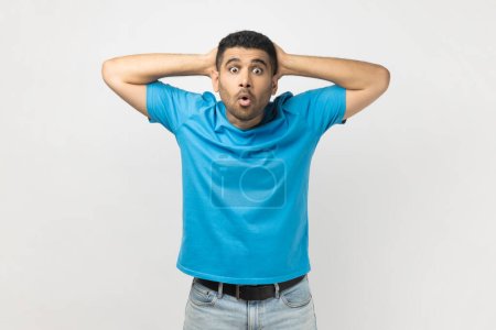 Portrait of shocked surprised man wearing blue T- shirt standing looking at camera with big eyes, keeps hands behind head, sees something astonished. Indoor studio shot isolated on gray background.