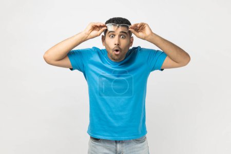 Photo for Portrait of shocked astonished unshaven man wearing blue T- shirt standing raised his optical eyeglasses, sees something surprised. Indoor studio shot isolated on gray background. - Royalty Free Image
