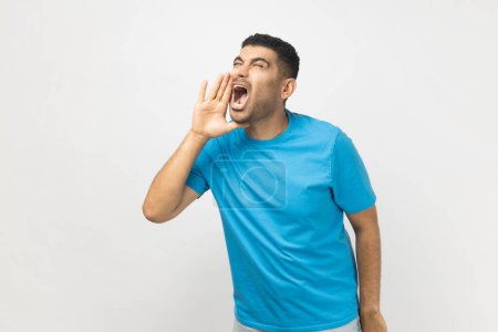 Photo for Portrait of excited handsome attractive unshaven man wearing blue T- shirt standing keeps hands near opened mouth, screaming news. Indoor studio shot isolated on gray background. - Royalty Free Image