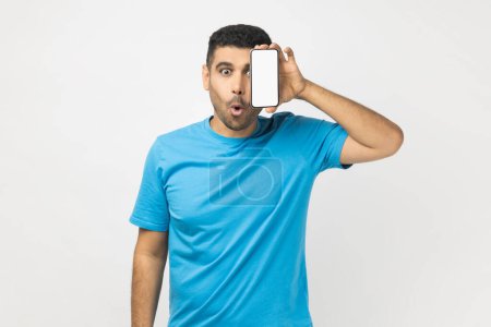 Téléchargez les photos : Surprised shocked astonished man wearing blue T- shirt standing covering his eye with mobile phone with empty display, advertisement area. Indoor studio shot isolated on gray background. - en image libre de droit
