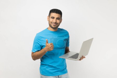 Téléchargez les photos : Portrait of happy delighted attractive unshaven man wearing blue T- shirt standing holding laptop in hands, working online, showing thumb up. Indoor studio shot isolated on gray background. - en image libre de droit