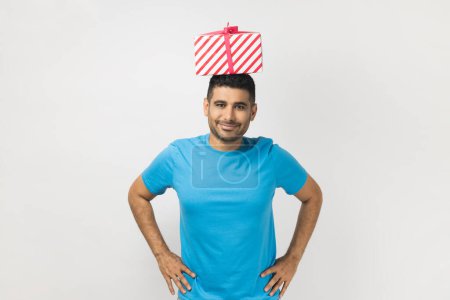 Photo for Portrait of satisfied delighted unshaven man wearing blue T- shirt standing with hands on hips, holding present box on his head. Indoor studio shot isolated on gray background. - Royalty Free Image