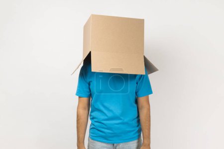 Photo for Portrait of unknown anonymous man wearing blue T- shirt standing with cardboard box on his head, having fun, hiding his face in carton parcel. Indoor studio shot isolated on gray background. - Royalty Free Image