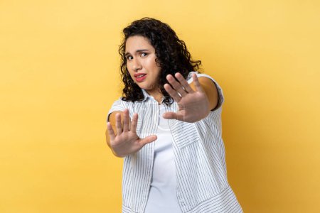 Foto de Portrait of scared beautiful woman with dark wavy hair gesturing stop with palms and looking surprised with frightened eyes. Indoor studio shot isolated on yellow background. - Imagen libre de derechos