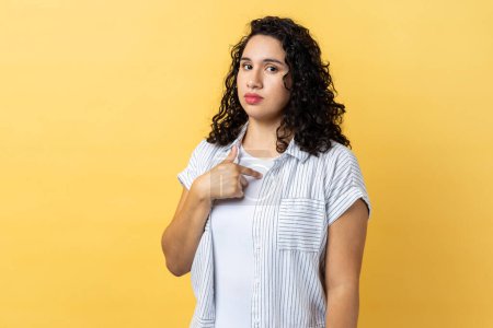 Foto de Portrait of serious self confident woman with dark wavy hair pointing at herself, feeling proud and self-important, having big ego. Indoor studio shot isolated on yellow background. - Imagen libre de derechos