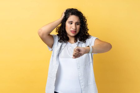 Photo for Portrait of woman with dark wavy hair standing with puzzled facial expression, worried about deadline, looking at wristwatch, keeps hand on head. Indoor studio shot isolated on yellow background. - Royalty Free Image