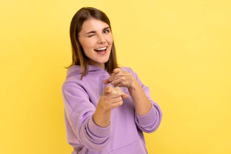 Foto de Happy satisfied woman pointing fingers on you looking at camera with toothy smile, congratulating with victory, wearing purple hoodie. Indoor studio shot isolated on yellow background. - Imagen libre de derechos