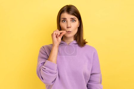 Téléchargez les photos : I don't tell anyone. Portrait of dark haired young adult woman standing, looking at camera and zipping mouth, wearing purple hoodie. Indoor studio shot isolated on yellow background. - en image libre de droit
