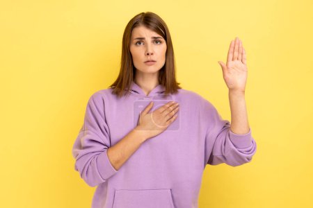 Téléchargez les photos : Serious conscious woman holding one hand on chest and raising another swearing, trust and honest, patriotism, wearing purple hoodie. Indoor studio shot isolated on yellow background. - en image libre de droit