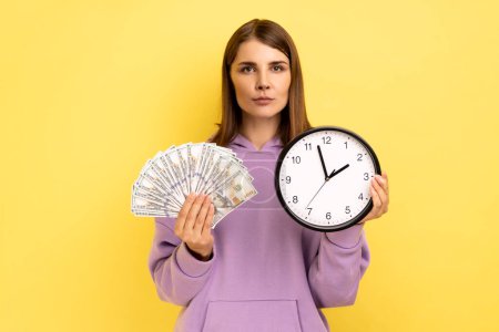 Téléchargez les photos : Rich dark haired woman holding big fan of dollar banknotes and wall clock, time is money, looking seriously at camera, wearing purple hoodie. Indoor studio shot isolated on yellow background - en image libre de droit