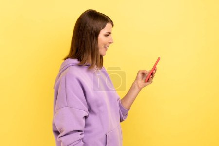 Téléchargez les photos : Side view of happy pretty woman reading message on smartphone and smiling, using mobile device for communication, browsing web, wearing purple hoodie. Indoor studio shot isolated on yellow background. - en image libre de droit