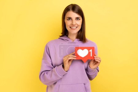 Foto de Cheerful carefree woman holding Heart Like icon of social media, notification button to follow, subscribe blog, popular forum, wearing purple hoodie. Indoor studio shot isolated on yellow background. - Imagen libre de derechos