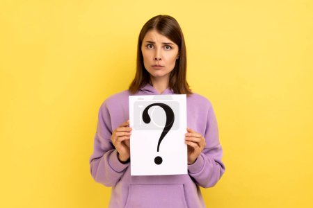 Téléchargez les photos : Portrait of serious concentrated woman looking at camera, holding paper with question mark, thinks about tasks, wearing purple hoodie. Indoor studio shot isolated on yellow background. - en image libre de droit