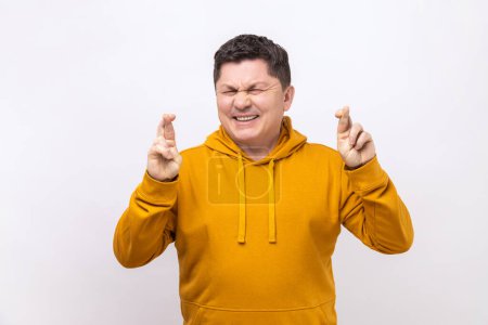 Foto de Portrait of middle aged man crossing fingers and closing eyes making wish, believing in miracle, hope in success, wearing urban style hoodie. Indoor studio shot isolated on white background. - Imagen libre de derechos
