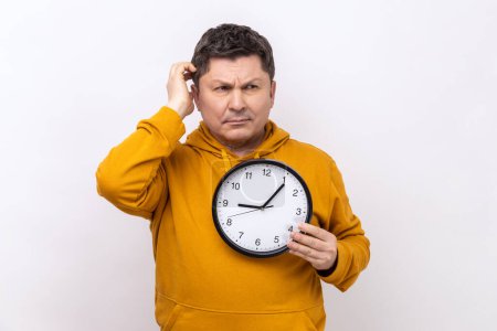 Photo for Portrait of middle aged man rubbing his head holding in hands big wallclock, has no time, worried about deadline, wearing urban style hoodie. Indoor studio shot isolated on white background. - Royalty Free Image
