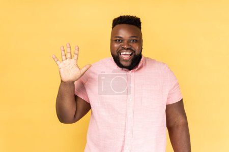 Foto de Portrait of friendly bearded man in pink shirt greeting you rising hand and waving, saying hi, glad to see you, looking at camera with toothy smile. Indoor studio shot isolated on yellow background. - Imagen libre de derechos