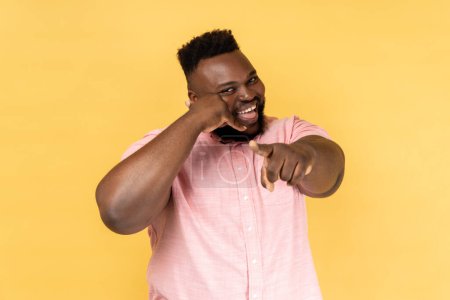 Foto de Portrait of smiling man wearing pink shirt holding fingers near ear showing telephone gesture looking and pointing at camera, waiting for your call. Indoor studio shot isolated on yellow background. - Imagen libre de derechos