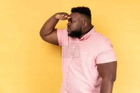 Foto de Side view of curious man with beard in pink shirt keeping palm over head and looking attentively far away, peering with expectation at long distance. Indoor studio shot isolated on yellow background. - Imagen libre de derechos
