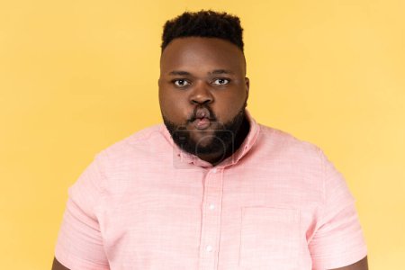 Foto de Portrait of funny positive young adult man wearing pink shirt standing with pouted lips making fish face, having fun, childish good mood. Indoor studio shot isolated on yellow background. - Imagen libre de derechos