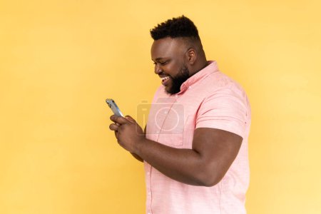 Téléchargez les photos : Side view of bearded happy man wearing pink shirt using mobile phone with happy expression, addicted to smartphone, texting in social network. Indoor studio shot isolated on yellow background. - en image libre de droit