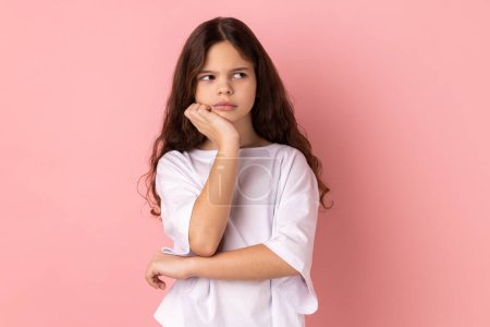 Foto de Portrait of thoughtful pensive little girl wearing white T-shirt thinking about future, holding chin, having serious facial expression. Indoor studio shot isolated on pink background. - Imagen libre de derechos