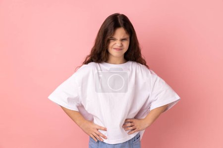 Foto de Angry aggressive little girl wearing white T-shirt standing with hands on hips, looking at camera with frowning face, expressing anger and hate. Indoor studio shot isolated on pink background. - Imagen libre de derechos