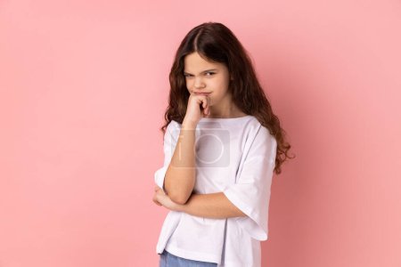 Photo for Need to think. Portrait of thoughtful little girl wearing white T-shirt holding her chin and pondering idea, confused not sure about solution. Indoor studio shot isolated on pink background. - Royalty Free Image