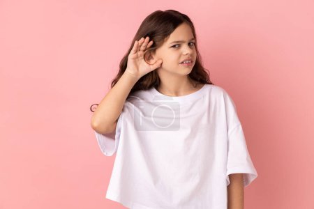 Téléchargez les photos : Portrait of curious little girl wearing white T-shirt holding hand near ear trying to hear secrets and find out gossips, listen to private whispers. Indoor studio shot isolated on pink background. - en image libre de droit