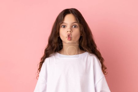 Foto de Portrait of excited funny little girl making fish face with lips and big amazed eyes, looking surprised and silly at camera, wondered expression. Indoor studio shot isolated on pink background. - Imagen libre de derechos