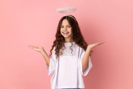 Foto de Portrait of smiling little girl wearing white T-shirt and with halo over head standing looking at camera spread hands aside, expressing happiness. Indoor studio shot isolated on pink background. - Imagen libre de derechos