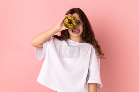 Téléchargez les photos : Portrait of joyful adorable little girl wearing white T-shirt standing having fun, holding and covering eye with donut and showing tongue out. Indoor studio shot isolated on pink background. - en image libre de droit