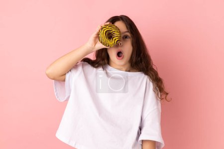 Photo for Portrait of excited amazed little girl wearing white T-shirt standing having fun, holding and covering eye with donut and keeps mouth open. Indoor studio shot isolated on pink background. - Royalty Free Image